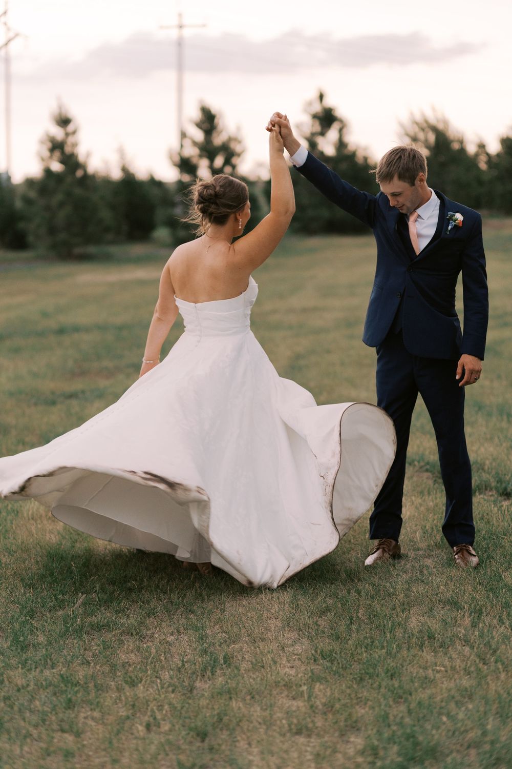 Groom in navy suit twirling bride in flowing white strapless wedding dress in green field at sunset.