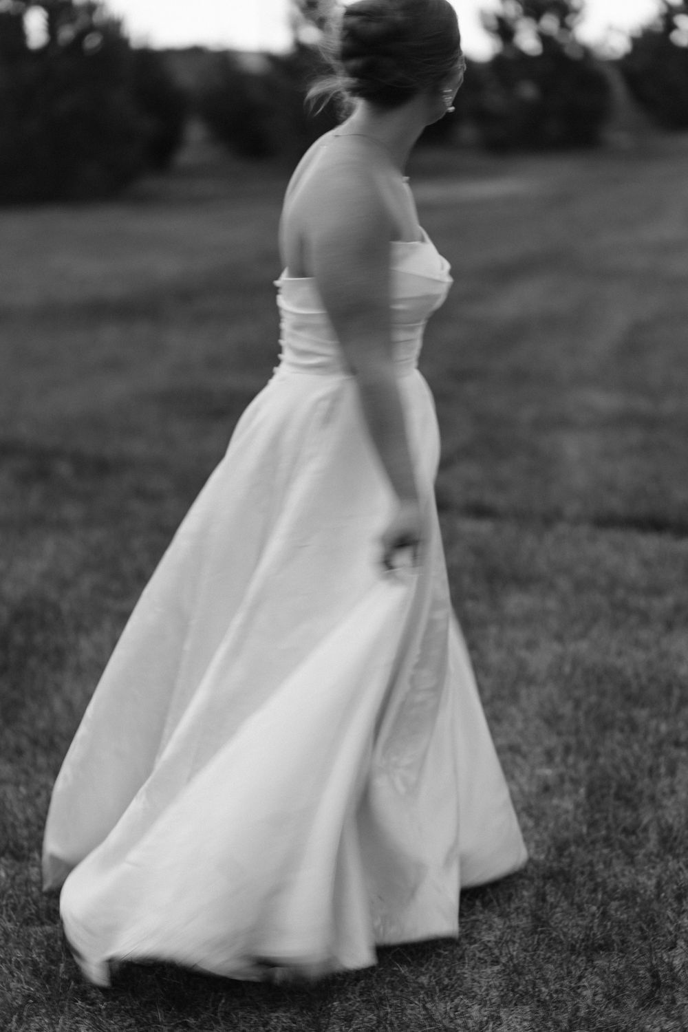 Black and white photo of bride in strapless ball gown walking gracefully across lush grass field.