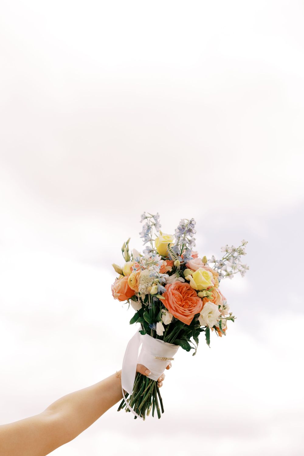 Hand holding a beautiful bridal bouquet of peach roses and blue flowers against a bright sky.