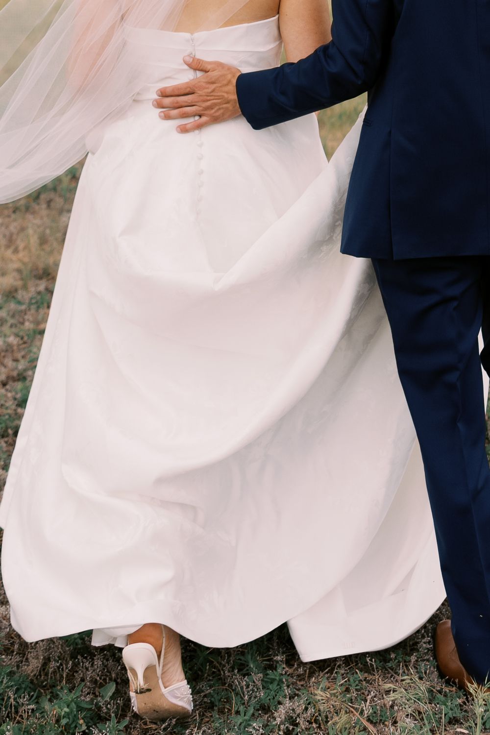 Groom in navy suit embracing bride in white wedding dress while walking outdoors together.