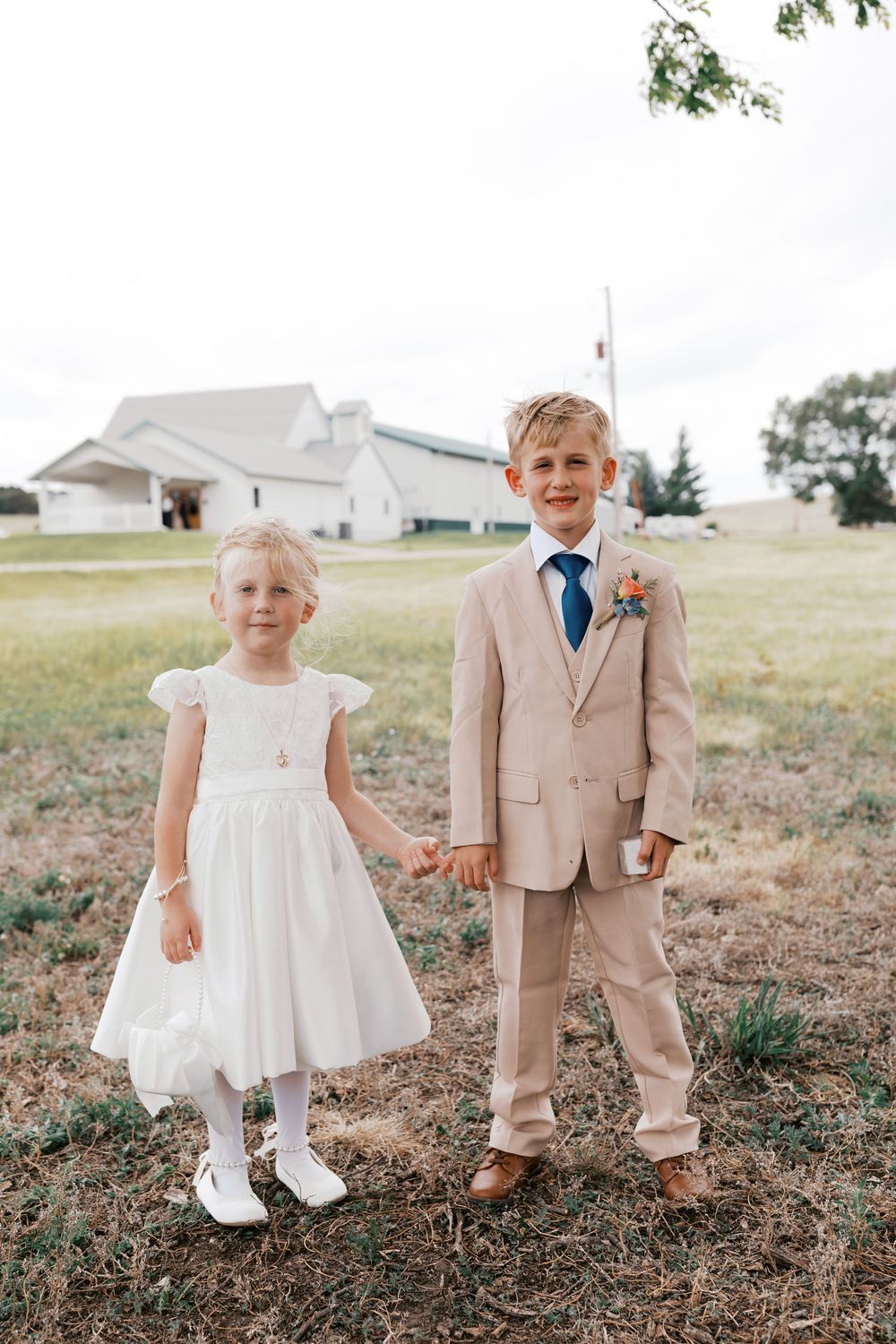 Adorable flower girl in white dress and ring bearer in tan suit hold hands at outdoor wedding.