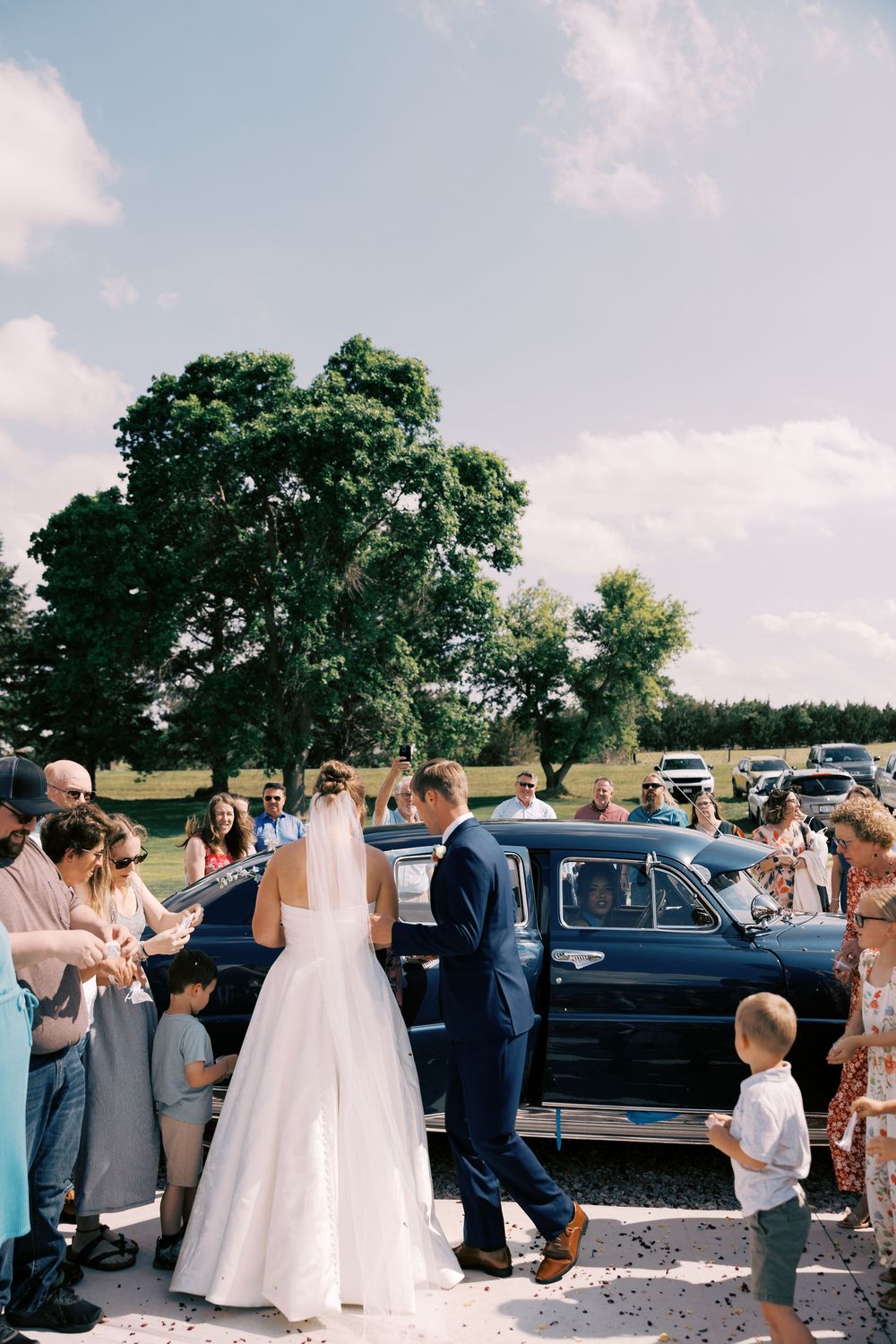 Bride and groom walk toward a vintage black car, greeted by guests throwing confetti on a sunny day.