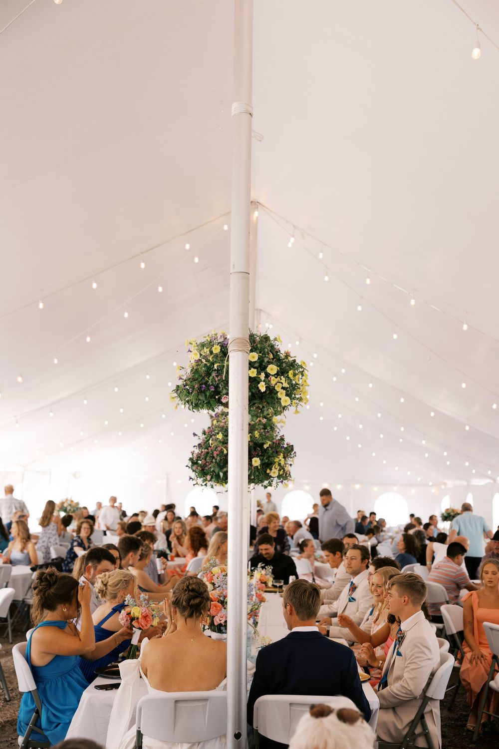 Guests celebrating inside a white wedding tent with string lights and hanging floral centerpiece above crowd.