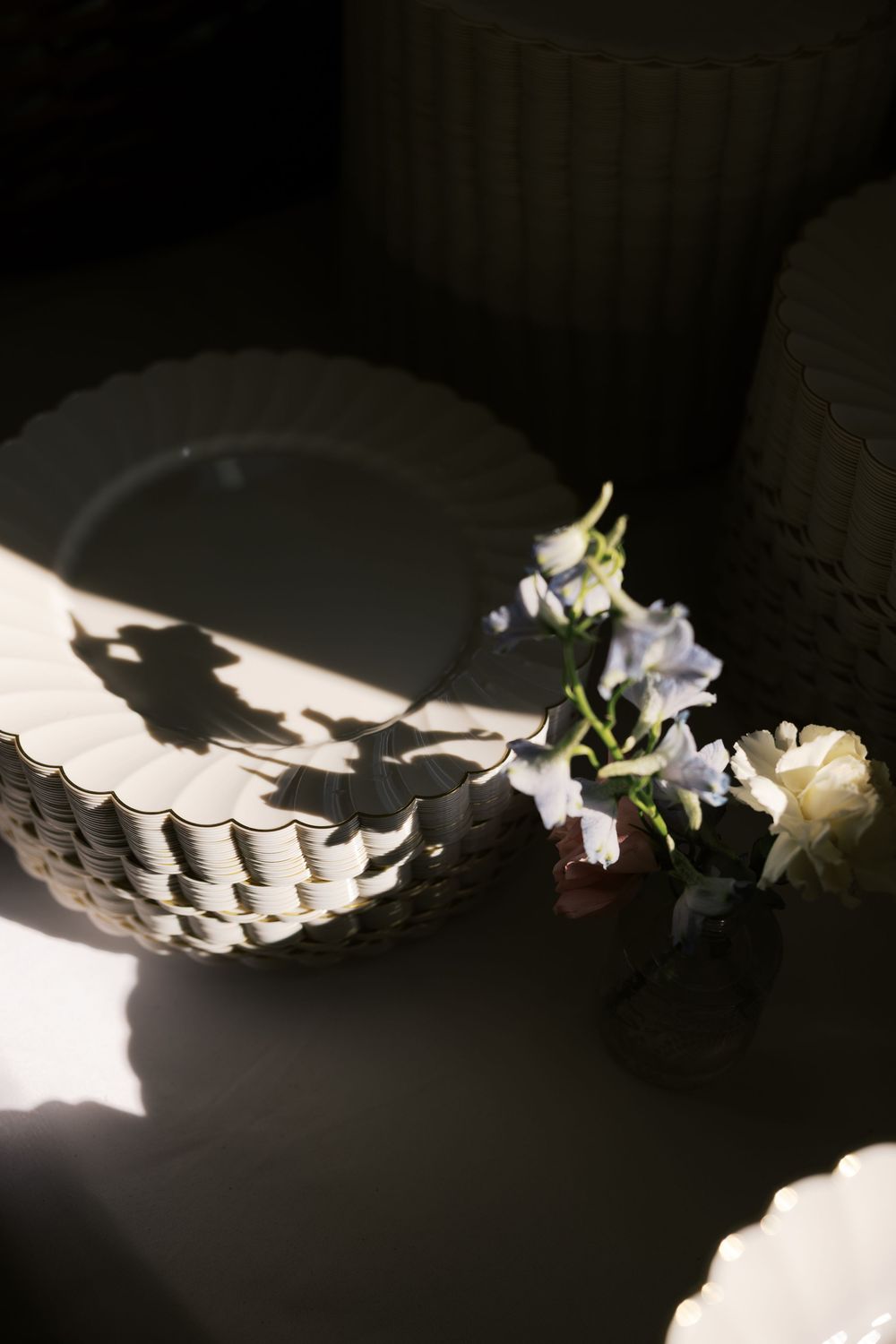 White scalloped plates and delicate blue flowers casting dramatic shadows in moody natural light on table.