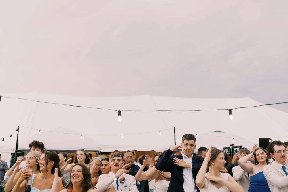 Joyful wedding guests dancing outdoors under string lights with white tent and overcast sky backdrop.