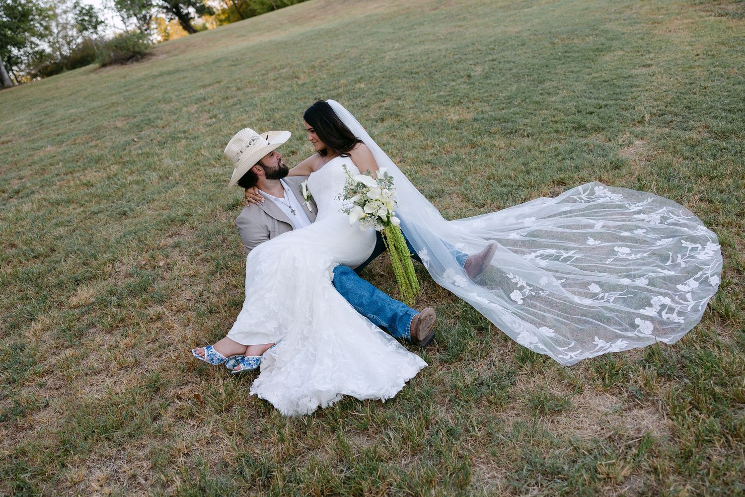 Bride and groom sitting on grass, bride in white gown with veil and bouquet, groom in jeans and cowboy hat.