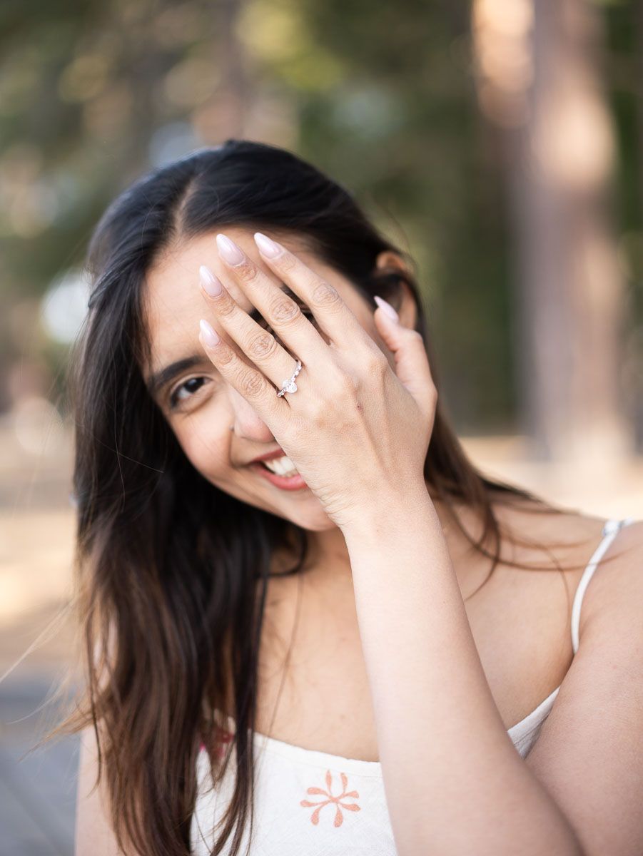 Young woman with long dark hair playfully covering her face with her hand, showing a delicate ring, smiling outdoors.