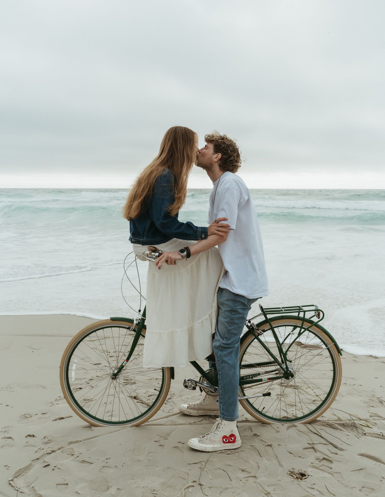Couple kissing on a green beach cruiser bicycle on a sandy beach with ocean waves in the background.