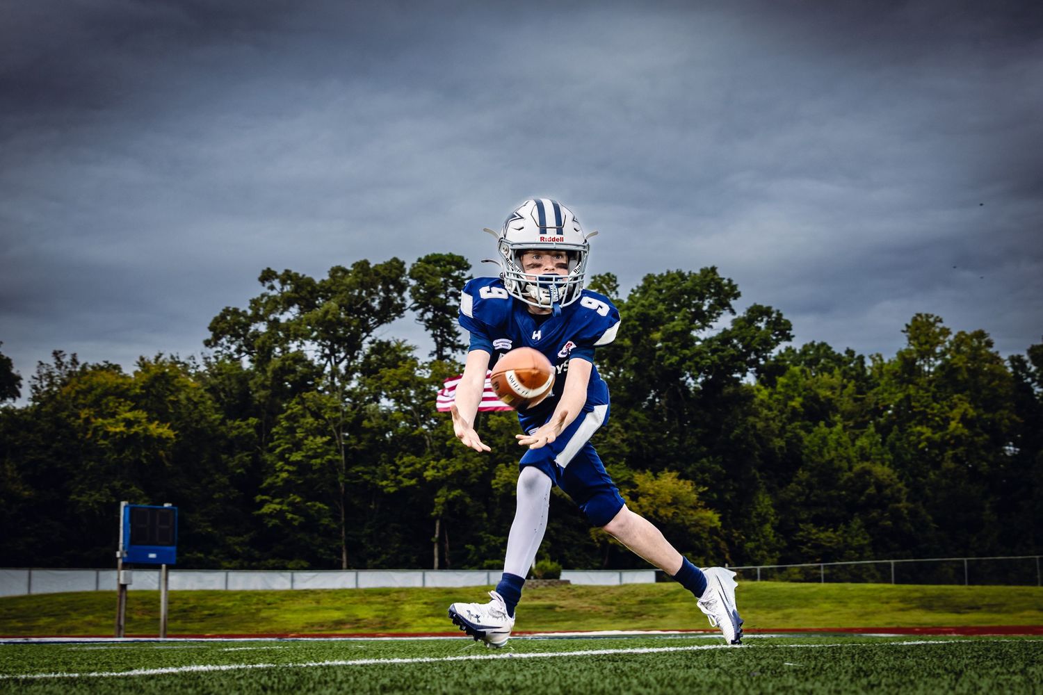 Young football player in blue uniform catches ball on field under dramatic cloudy sky.
