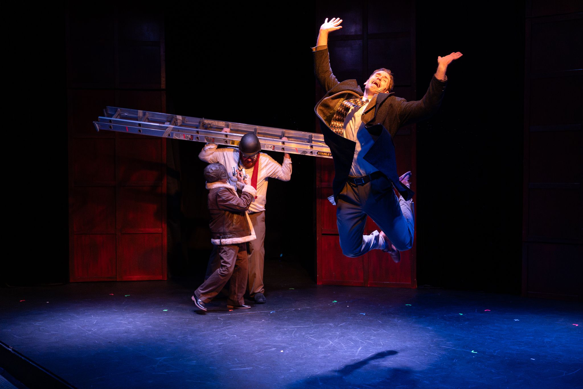 Two performers on a dark stage, one leaping dramatically in the air while another crouches near a ladder.
