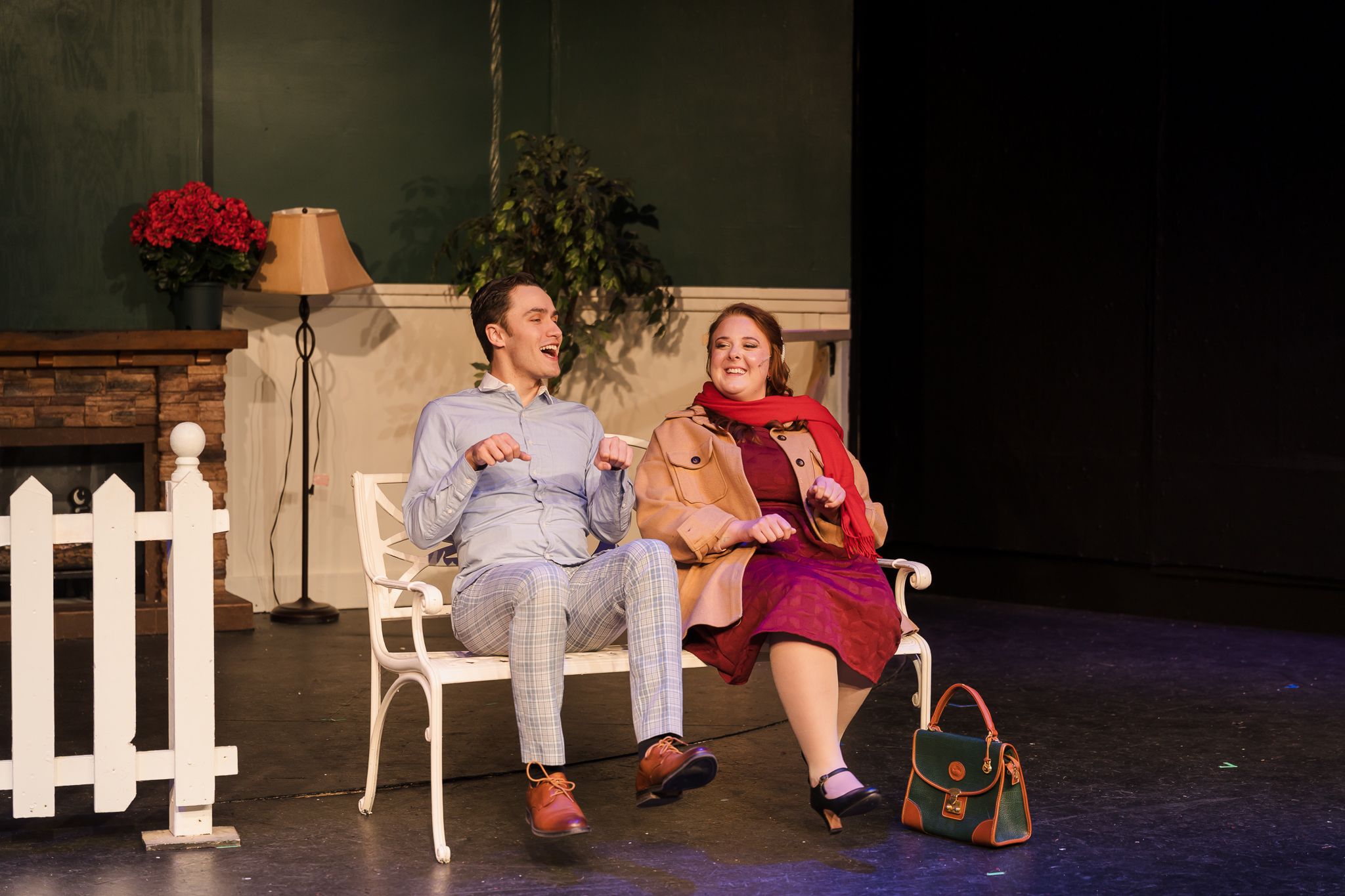 Two actors perform a theater scene, sitting on a white bench on stage with props including a lamp and handbag.