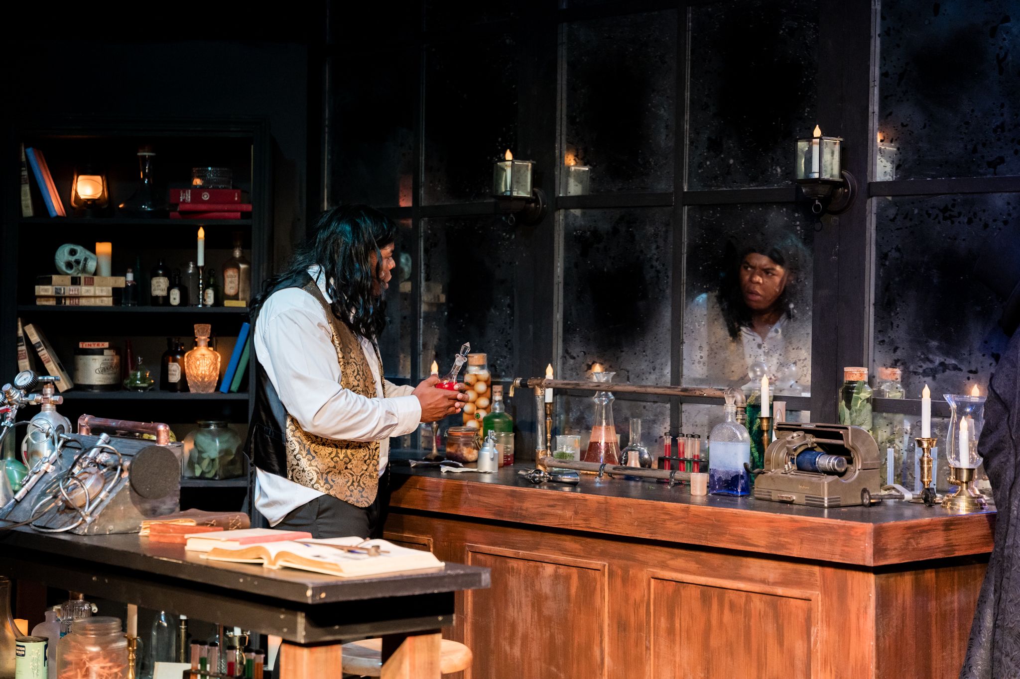 Two actors perform in a candlelit theatrical set resembling a Victorian apothecary shop with shelves of bottles.