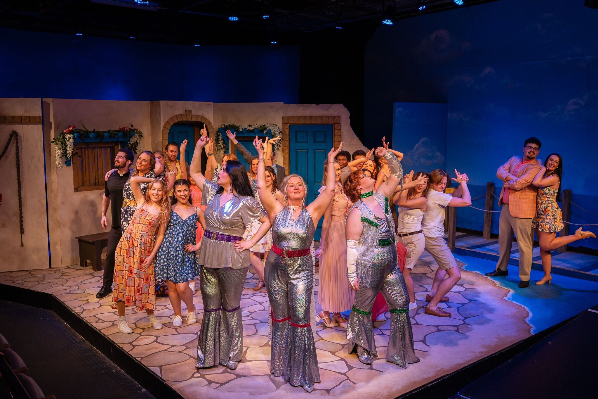 A colorful ensemble cast performs a lively musical theater production on a decorated stage with blue backdrop lighting.