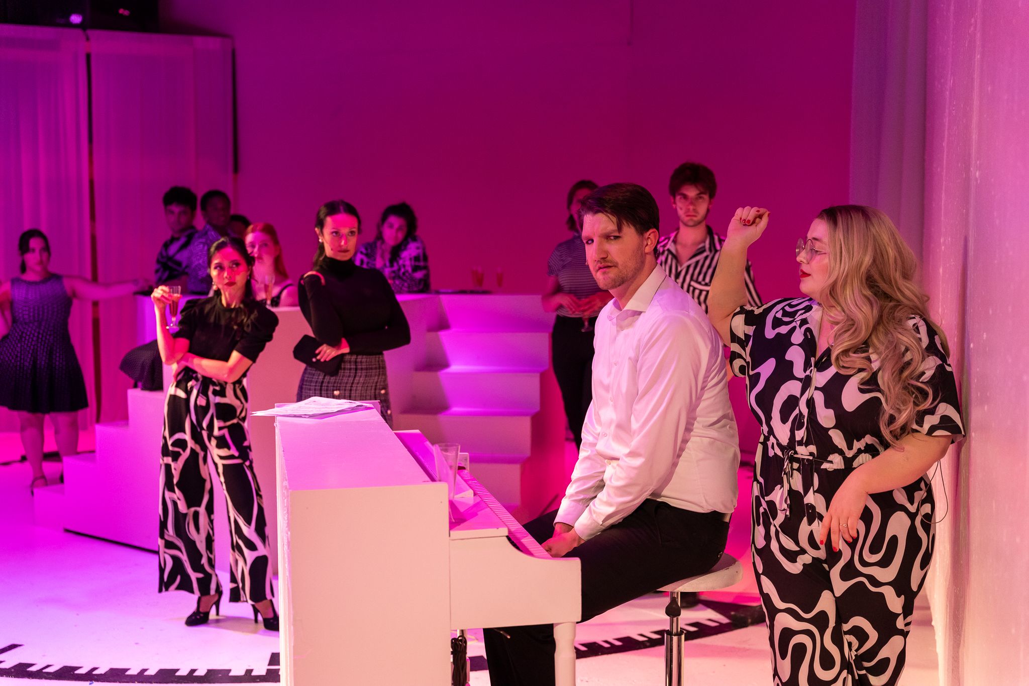Man playing white piano at elegant pink-lit party with guests in black and white outfits in background.