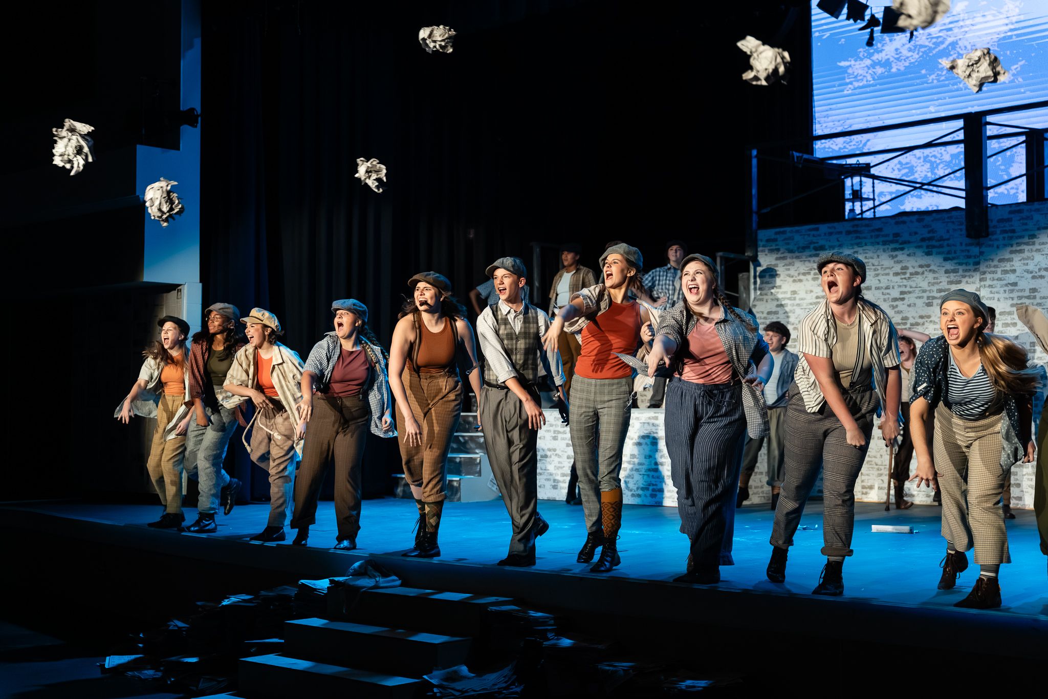 Theater performers in period costumes sing and dance on a dramatic stage with blue lighting and scenic backdrop.