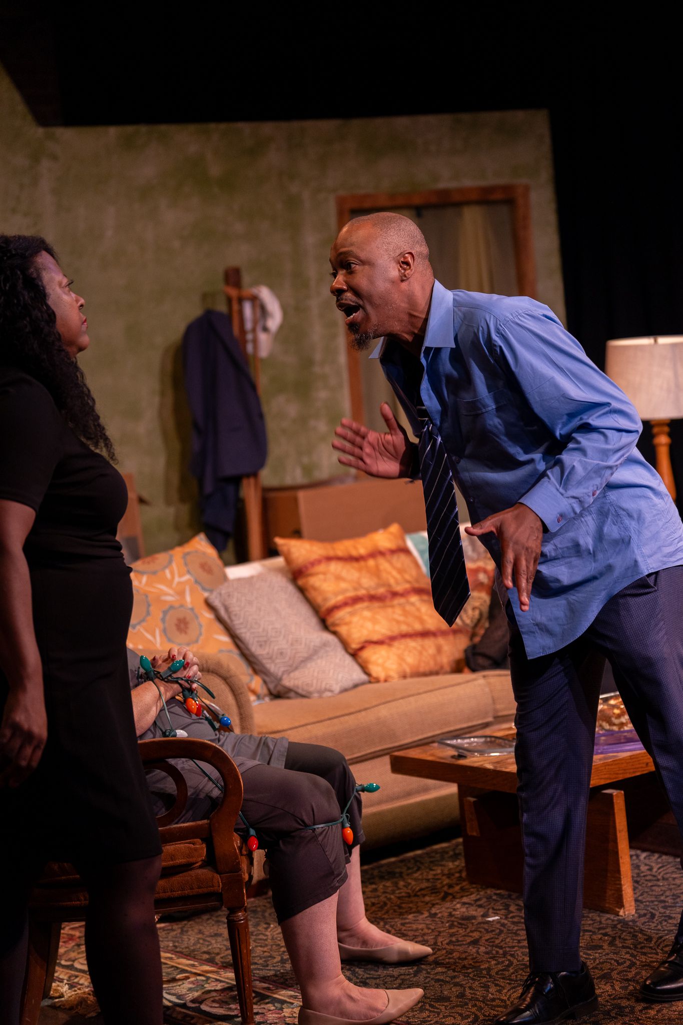 Two actors perform an intense dramatic scene in a theater, with a man in a blue shirt confronting a seated woman on stage.