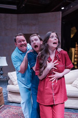 Three actors in pajamas perform a comedic theater scene, expressing exaggerated shock and surprise on a living room stage set.