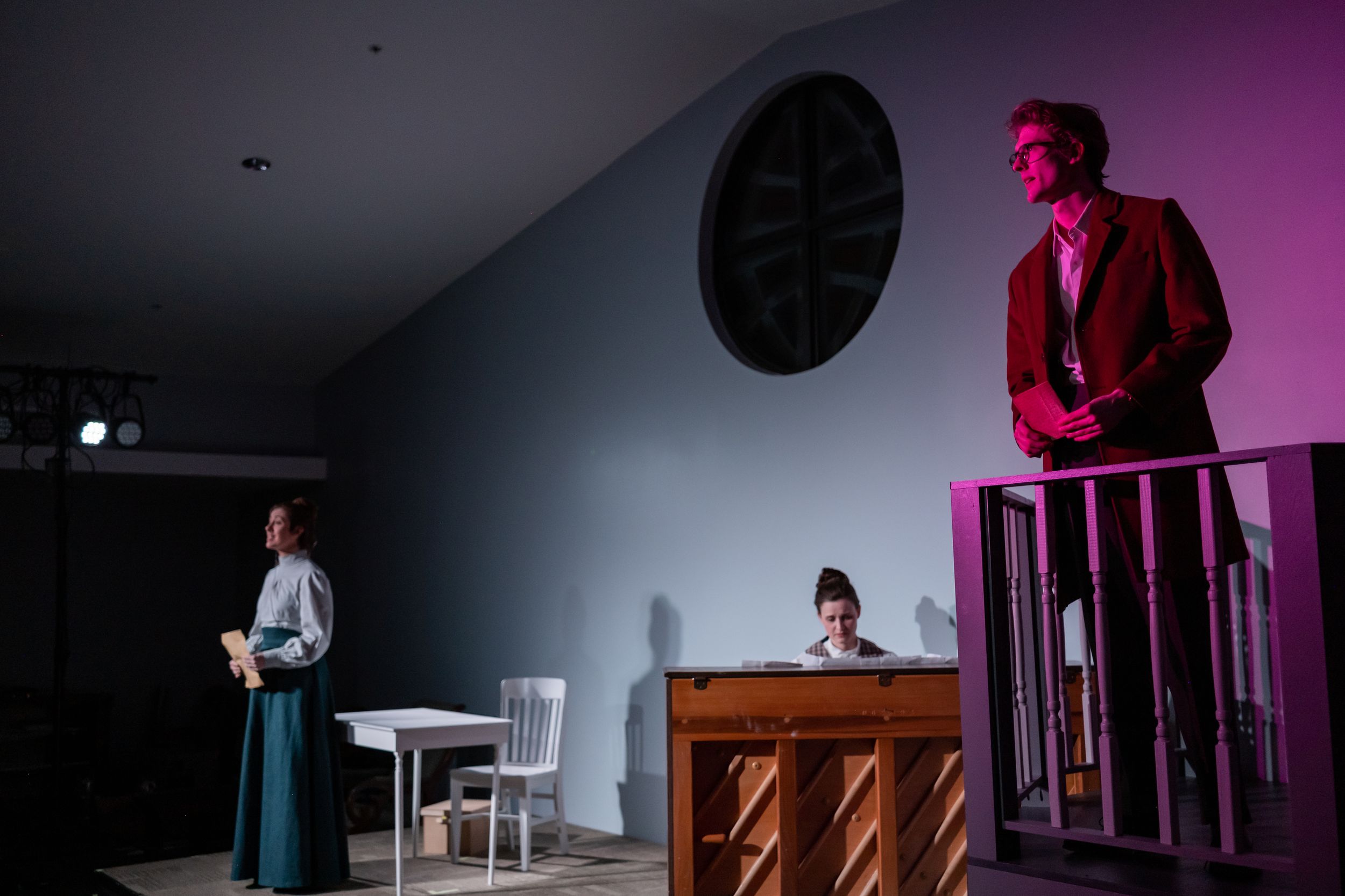 Three actors perform on a dramatic stage set with moody pink and blue lighting during a theater production.