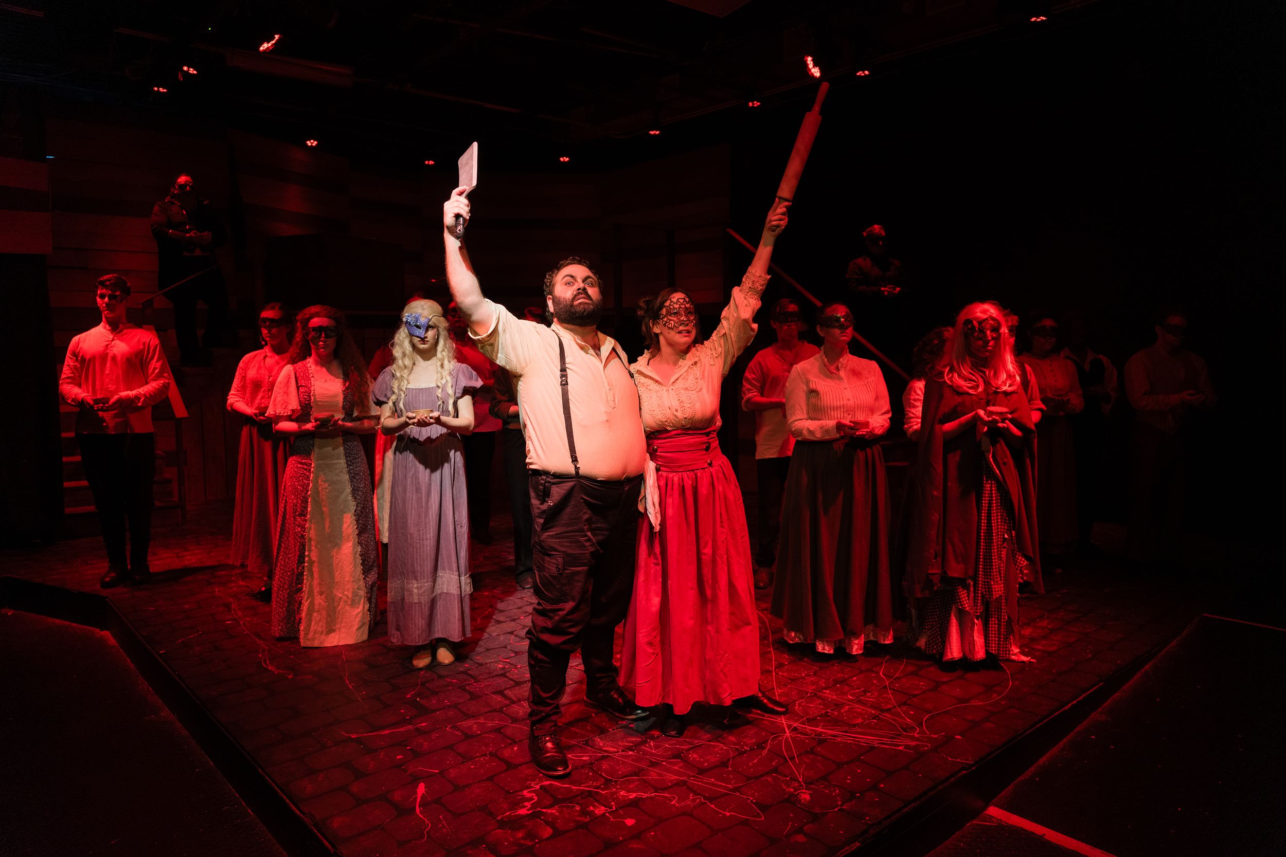 A bearded actor raises his arms dramatically on a red-lit stage surrounded by ensemble cast members in period costumes.