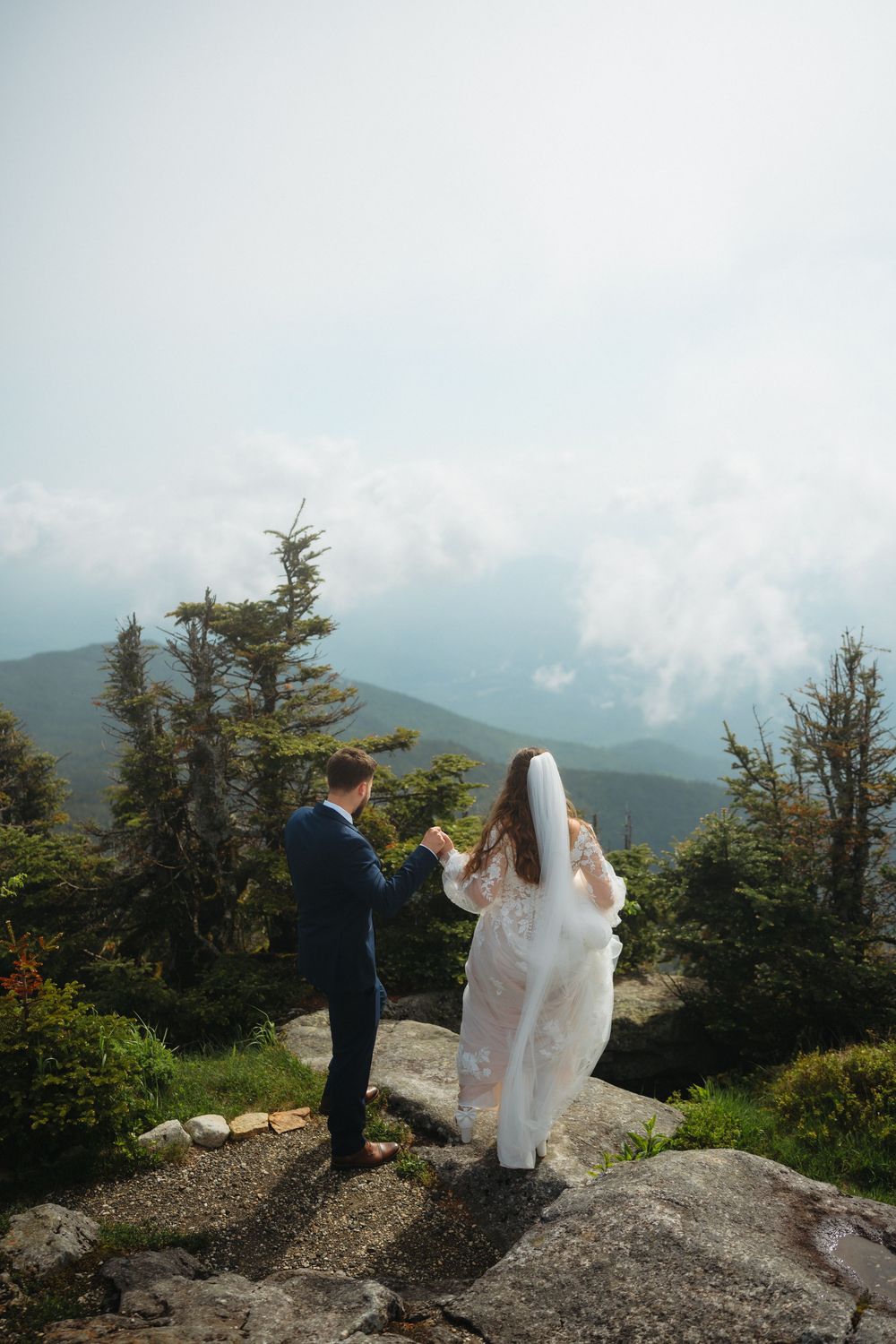 Bride and groom stand on rocky mountain summit holding hands with misty peaks and evergreen trees in background.
