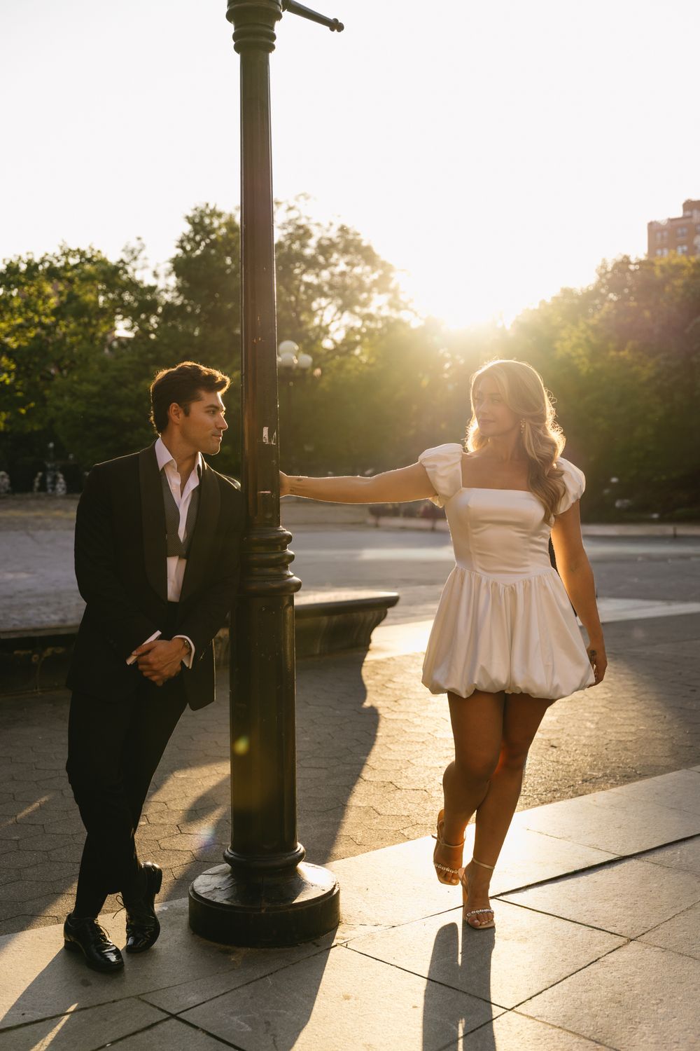 Couple in elegant attire sharing a romantic moment by a lamp post at golden hour in an urban park.