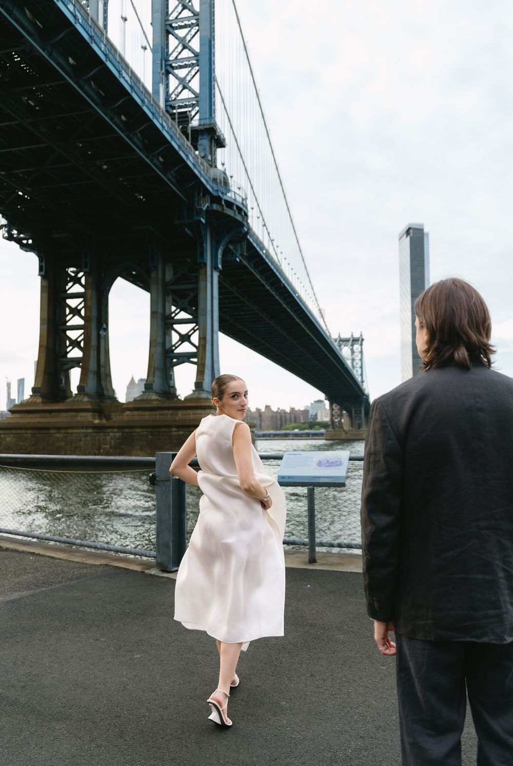 Woman in white dress walks near Manhattan Bridge waterfront as man in black suit watches.