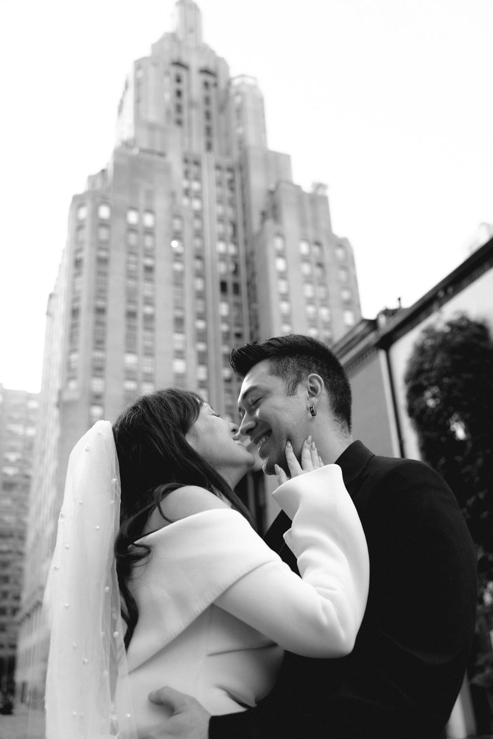 Black and white photo of a couple nearly kissing in front of a tall Art Deco skyscraper, bride in white veil.