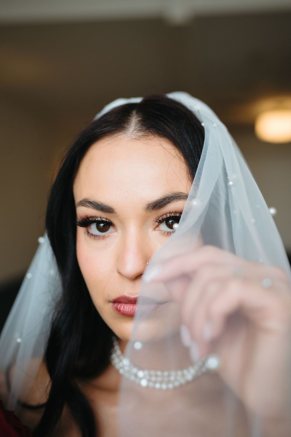 A bride in a white pearl-trimmed veil and pearl necklace gazes at the camera, holding her veil delicately.