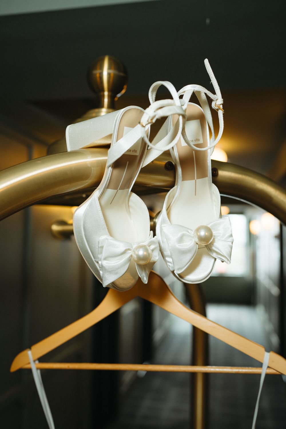 White bridal heels with pearl bow detail hanging on a gold luggage rack, perfect wedding day shoes.