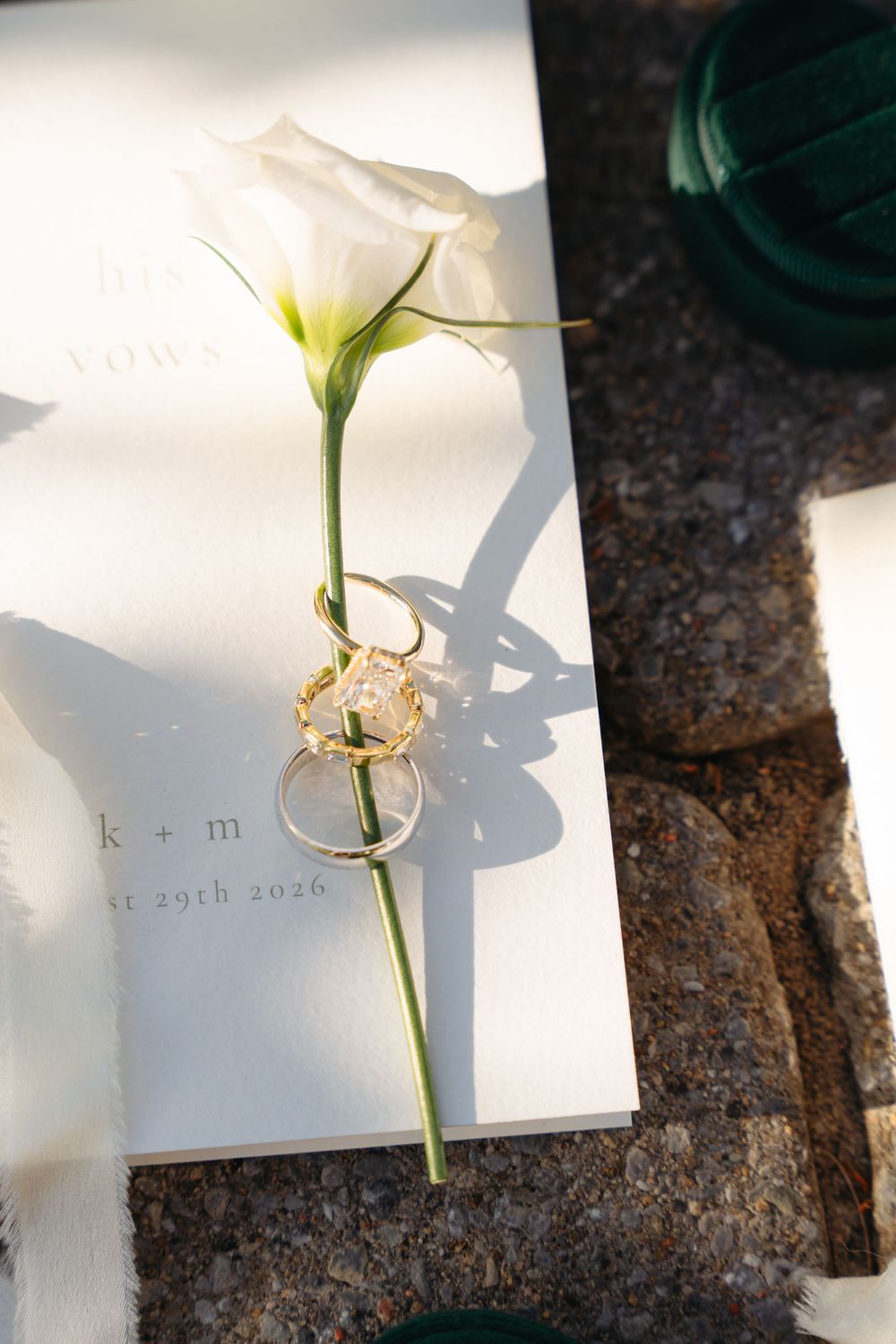 Wedding vows booklet with white calla lily and gold engagement ring with wedding band on stone surface in sunlight.