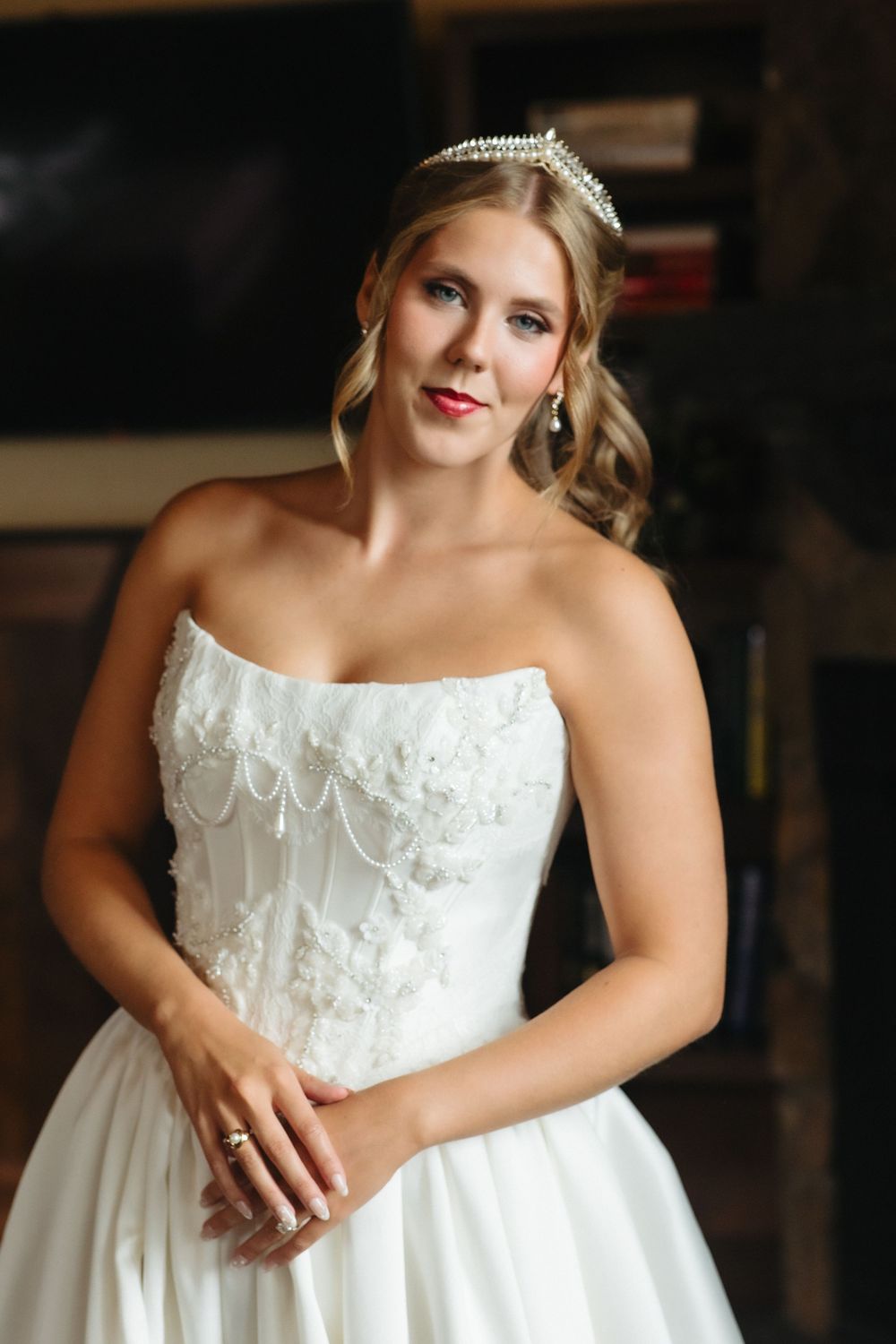Elegant bride in strapless white lace wedding gown wearing pearl tiara and drop earrings, posing indoors.