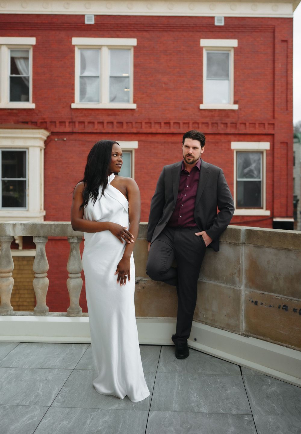 Elegant couple posing on rooftop, woman in white gown and man in dark suit, red brick building backdrop.