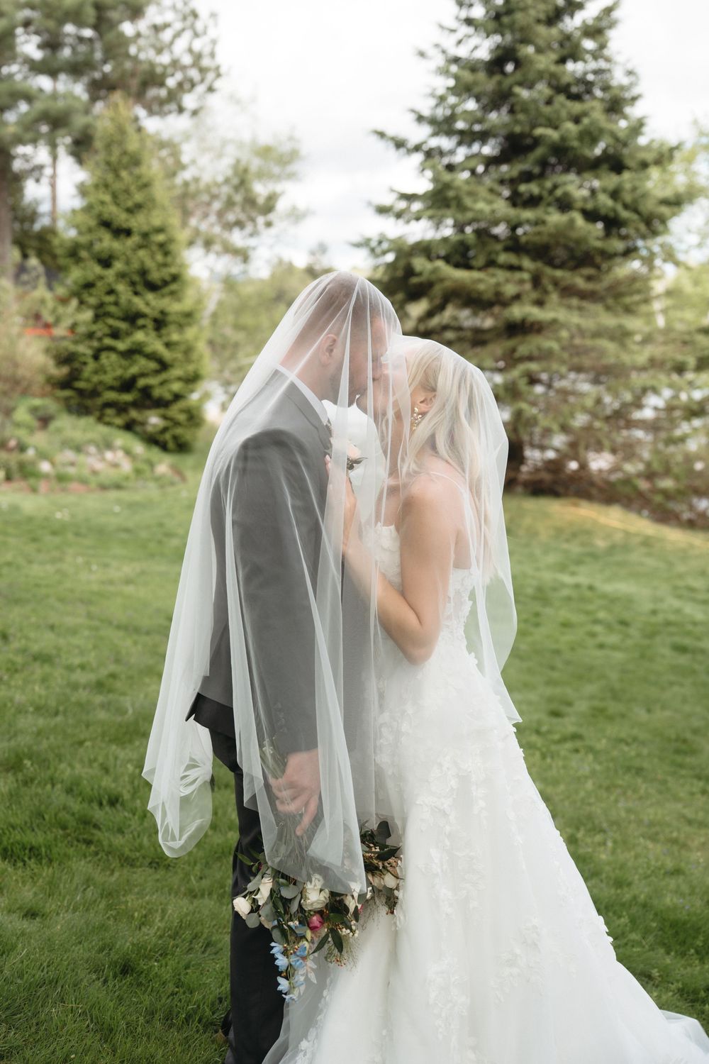Bride and groom kissing under a white veil outdoors, surrounded by lush green trees, bride holding a floral bouquet.