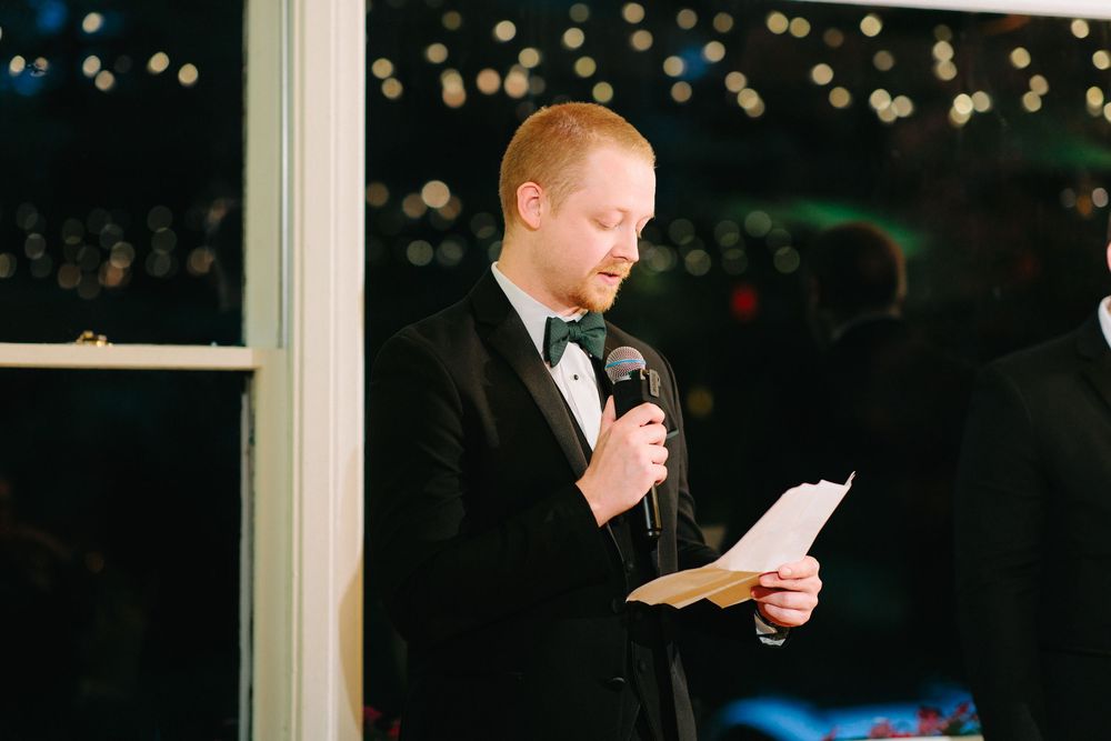 A man in a black tuxedo reads a speech into a microphone at an elegant evening wedding reception.