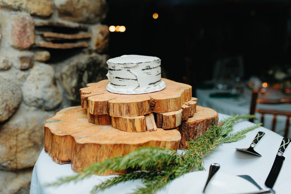 Rustic wedding cake displayed on stacked wooden log slices with evergreen greenery on white tablecloth.