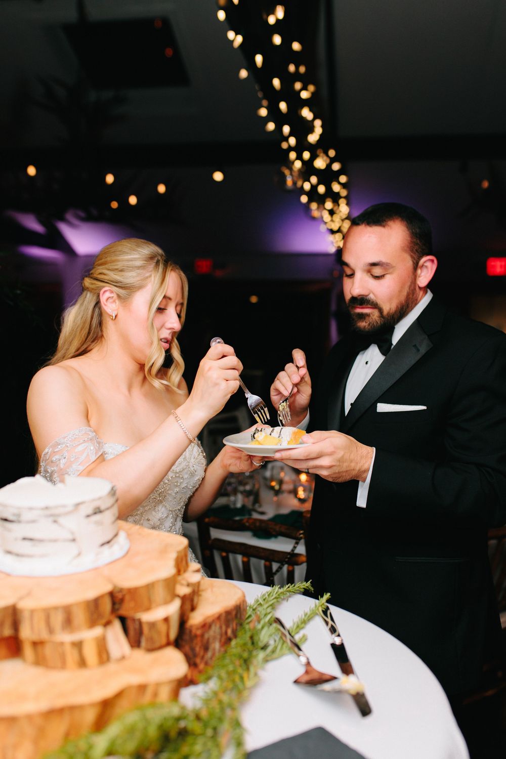 Bride and groom share first bite of rustic wedding cake displayed on wooden log slices.