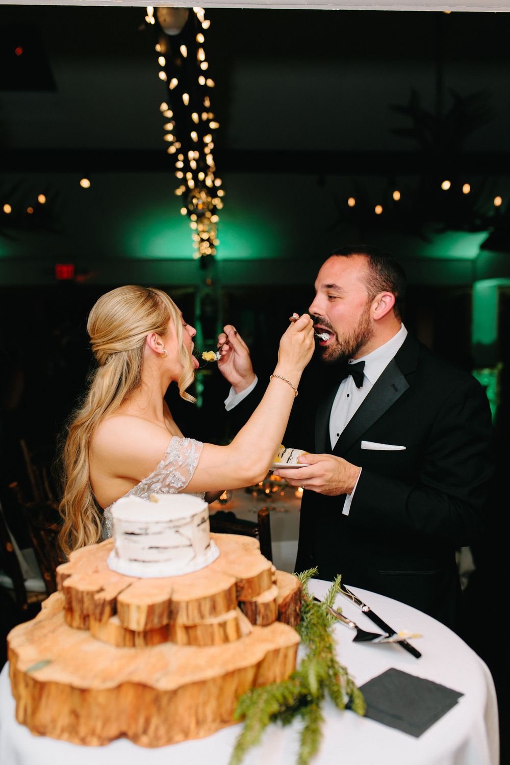 Bride and groom feeding each other wedding cake at rustic reception with string lights and wood slice cake stand.