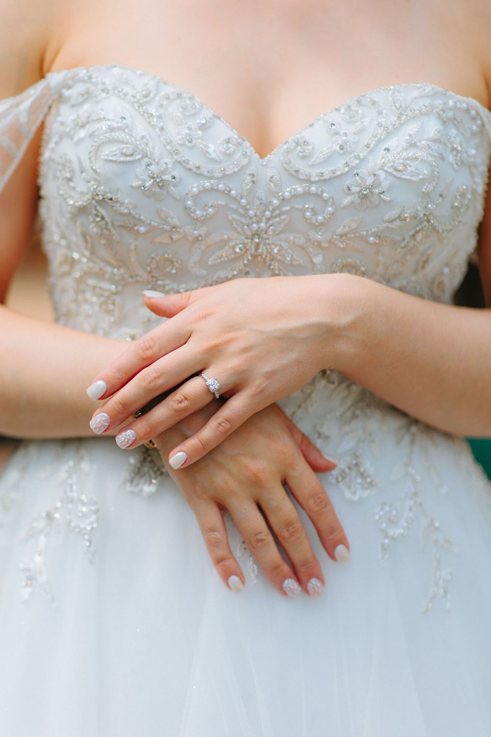 Close-up of bride's hands folded over intricate beaded white wedding gown bodice with diamond ring.