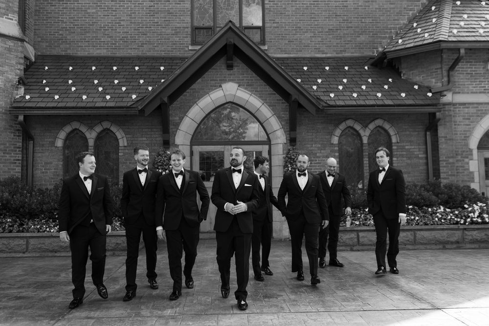 Black and white photo of groom and groomsmen in tuxedos posing outside elegant brick church.