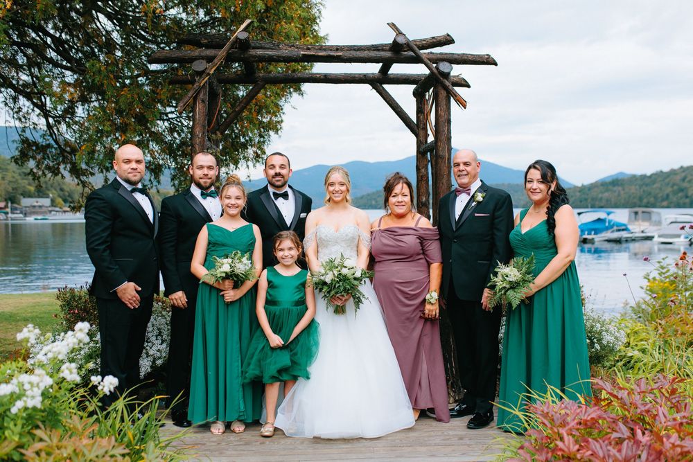Wedding party in emerald green and mauve dresses with groom in tuxedo posing under rustic wooden arch by a mountain lake.