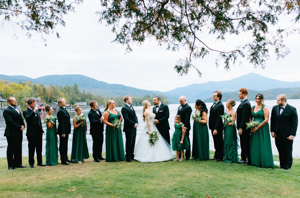 Wedding party in emerald green dresses and black tuxedos posing by a lake with mountain views.