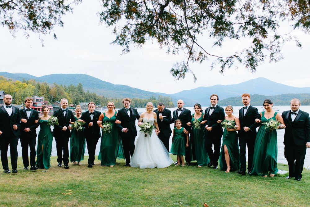 Large wedding party in black tuxedos and emerald green gowns pose by a mountain lake.