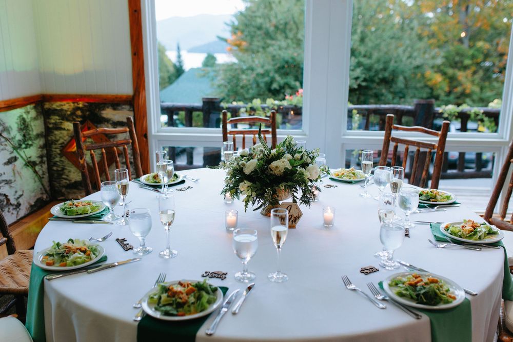 Wedding reception round table with white linen, green napkins, floral centerpiece, and scenic outdoor view through windows.