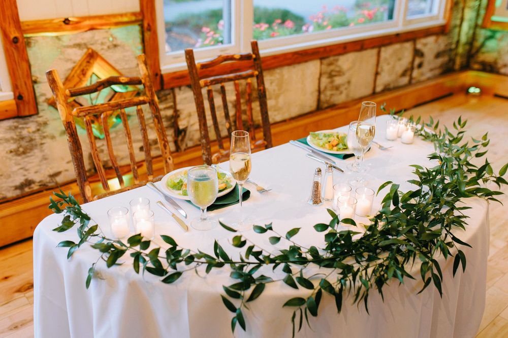 Romantic sweetheart table with white linen, greenery garland, candles, and wooden chairs for wedding reception.