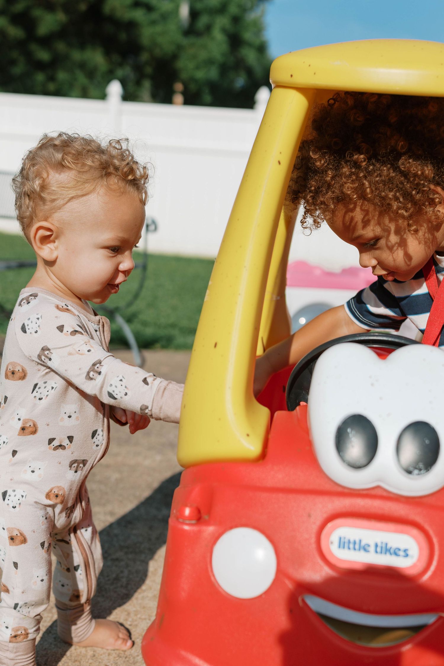 Two young toddlers play outdoors with a red and yellow Little Tikes Cozy Coupe toy car.