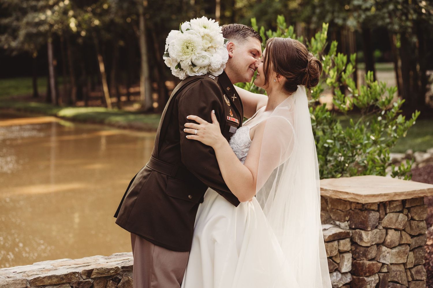 A bride and groom share a romantic moment outdoors near a pond, with lush greenery in the background.