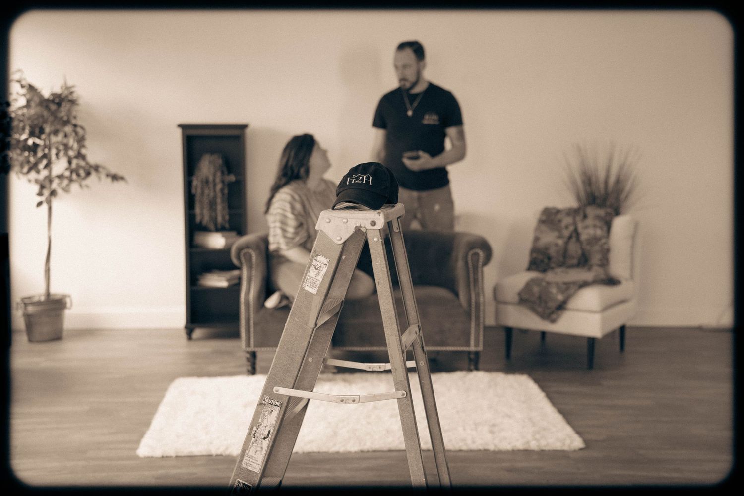 Woman sitting on a ladder while a man stands nearby in a cozy living room with vintage furniture.