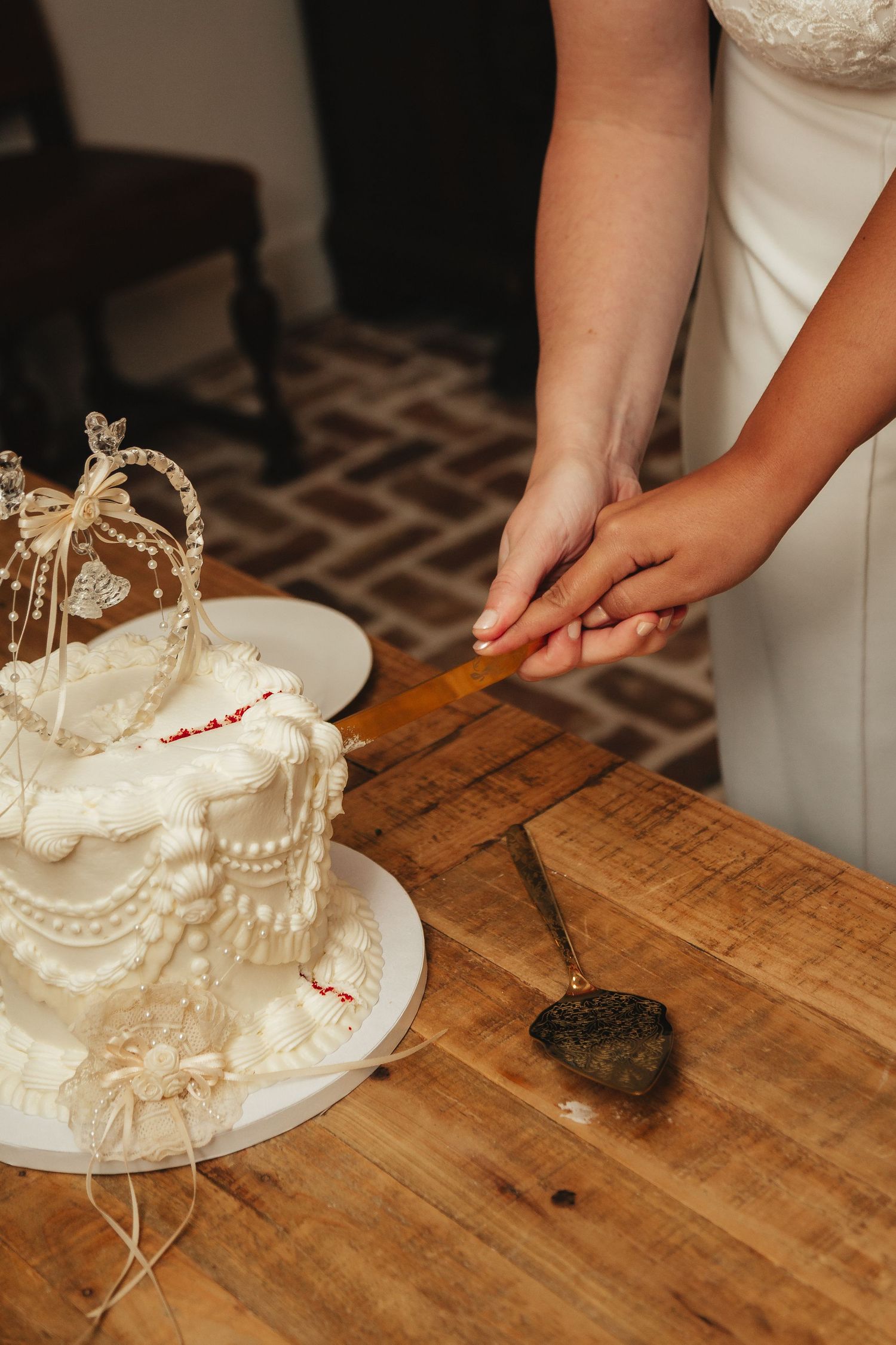 Bride cutting a white wedding cake topped with a crown decoration on a wooden table with a heart-shaped cake server.