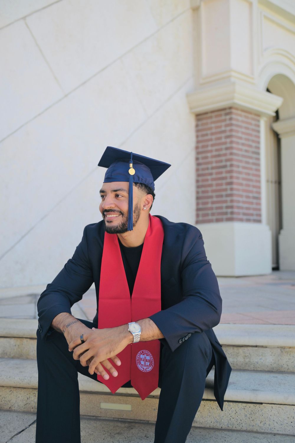 Smiling male graduate sitting on steps wearing a navy cap and red sash over a dark suit, celebrating his achievement.