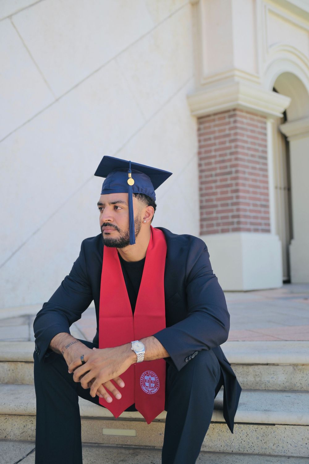Young man in graduation cap and red sash sits on steps outside university building in formal suit.